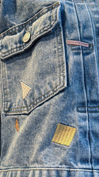 istock Full frame image of denim jacket displaying buttoned down pocket with embroidered shapes in store, metal button, sewn square and triangle design, focus on foreground 2117224359