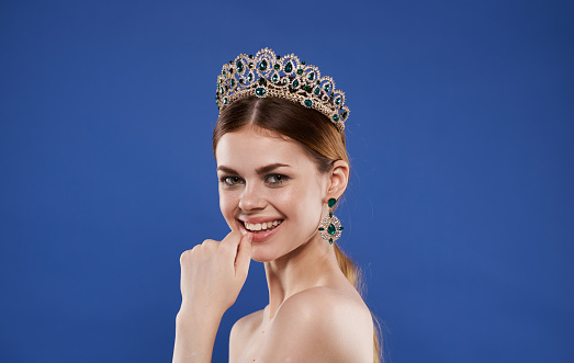 A woman with bare shoulders and a diadem on her head gestures with her hands on a blue background. High quality photo