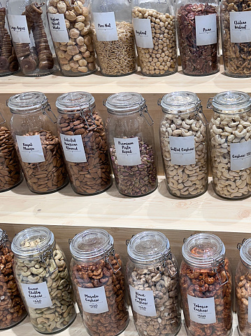 Stock photo showing whole food store pantry with an assortment of glass jars full of various dried ingredients, shelled and seasoned nuts including pine nuts, hazelnuts, pecans, walnuts, almonds, pistachios and cashews which have been flavoured with tabasco, green chillies, masala, salt or black pepper. These jars are lined up and organised neatly on shelves for ease of access, with labels on them.