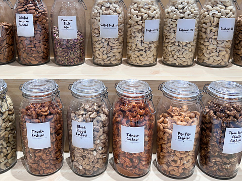 Stock photo showing whole food store pantry with an assortment of glass jars full of various dried ingredients, shelled and seasoned nuts including pecans, almonds, pistachios and cashews which have been flavoured with tabasco, green chillies, masala, salt or black pepper. These jars are lined up and organised neatly on shelves for ease of access, with labels on them.