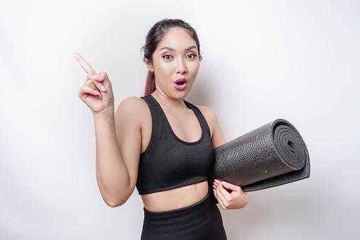 Shocked sporty Asian woman wearing sportswear is holding yoga mat on isolated white background.