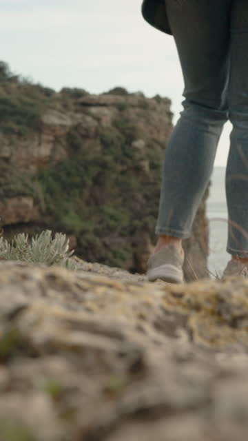 Vertical video. Close-Up, Focused on the Legs and Sneakers of a Woman Walking on Rocks towards the Edge of a Cliff by the Sea. The Focus Follows the Feet.