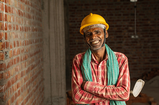 Portrait of smiling senior male mason with arms crossed holding trowel and standing by brick wall at construction site
