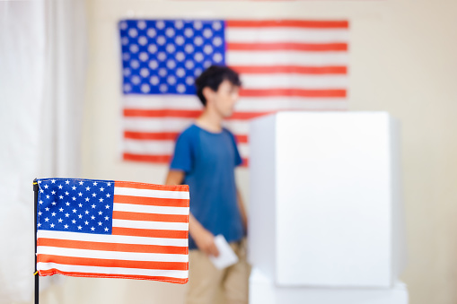 A man casts his vote in a voting booth to elect the President of the United States. A small US flag decorates the voting area in the foreground