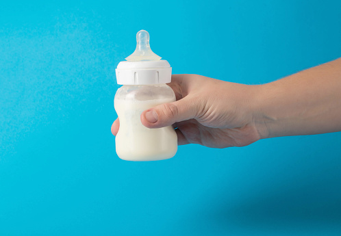Father's hand with a baby bottle with milk formula on a blue background shakes the mixture to mix. Nutrition of young children for health. Breast milk oligasacchorides. Copy space for text, porridge