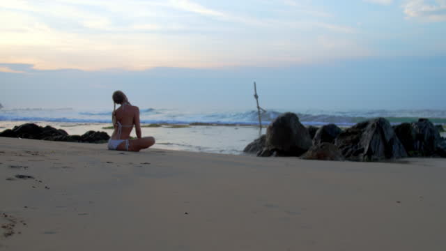 Woman silhouette in bikini meditating on beach by rocks