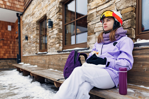 Young woman skiing in the Alps mountains. She is taking a break to eat sandwich and drink hot tea from insulated drink container. 
Overcast winter day.
Shot with Canon R5.