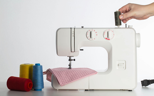 The hand of a seamstress holds a modern automatic sewing machine on a white background with multi-colored spools of thread. Copy space for text