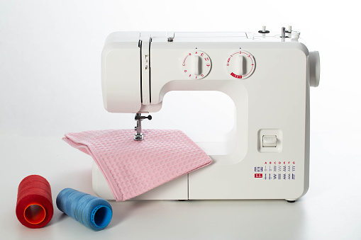 Modern automatic white sewing machine with pink fabric and multi-colored spools of thread on the table. White background. Tailoring and clothing repair services.