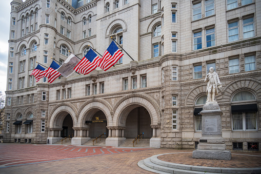 Washington DC, USA - March 19, 2024: Entrance to the Waldorf Astoria Hotel in Washington DC. A row of American flags is over the arched entrance. A statue of Ben Franklin is in front.