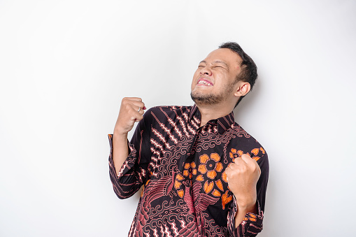 A young Asian man with a happy successful expression wearing batik shirt isolated by white background