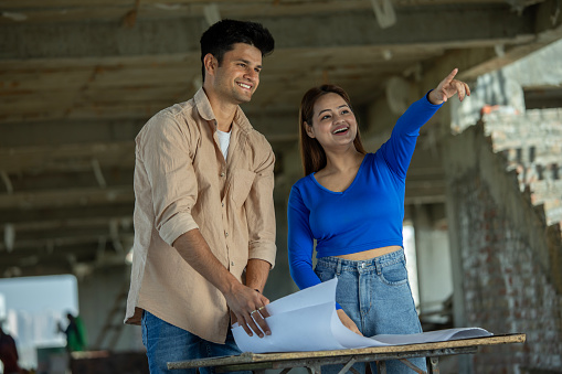 Smiling young couple analyzing and discussing apartment blueprint on table while standing in construction building