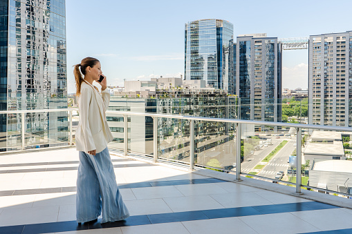 A woman is walking on a balcony overlooking a financial district while talking on her cell phone