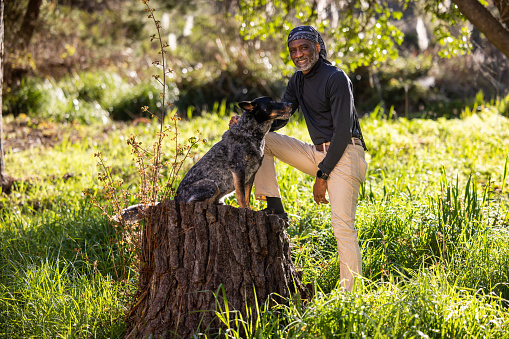 Portrait of black man in the park with his well-trained Blue Heeler, Australian Cattle Dog  sitting on a log enjoying each other's company on a bright and sunny day.