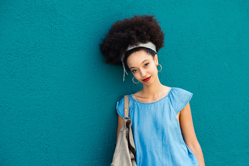 Portrait of smiling young Moroccan female in makeup with headband afro hair looking at camera while standing with handbag against blue background in daylight