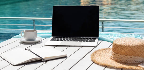 istock Alternative workplace at resort poolside. Laptop, notepad, cup of coffee and hat on white table isolated on blue background of pool water. Blank computer screen, copy space for text or design. Banner 2117616094