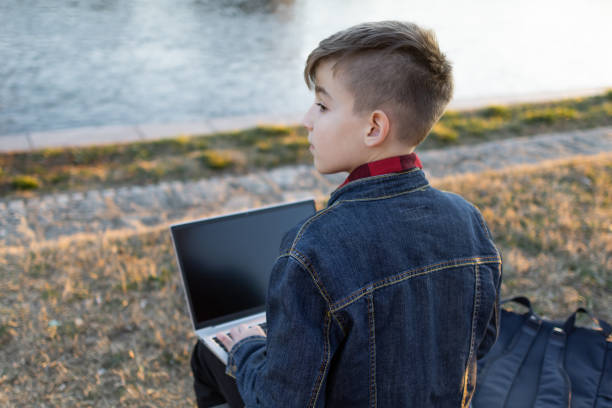 Cute boy is sitting on a bench near the river at sunset and using laptop to communicate with friends. Online education.