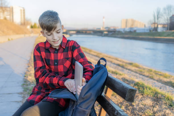 istock Cute boy is sitting on a bench near the river at sunset and using laptop to communicate with friends. Online education. 2117648054
