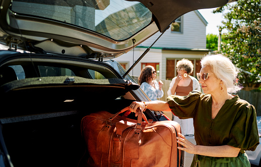 Mature woman packing bags into the trunk of a car in a driveway while preparing for a weekend getaway with a group of female friends