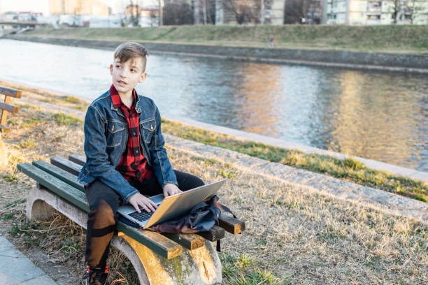istock Cute boy is sitting on a bench near the river at sunset and using laptop to communicate with friends. Online education. 2117663553