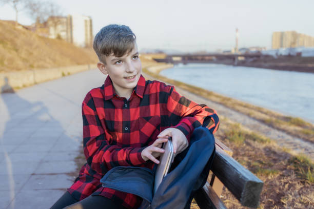 Cute boy is sitting on a bench near the river at sunset and using laptop to communicate with friends. Online education.