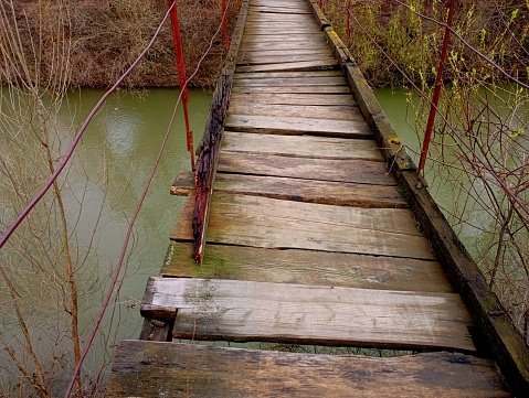 Old wooden bridge with broken and rotten planks underfoot across the river. Pedestrian dangerous emergency bridge to the opposite bank of the river.