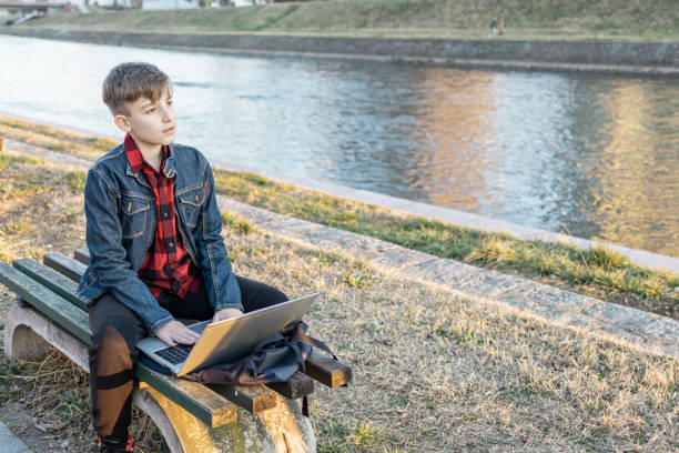 istock Cute boy is sitting on a bench near the river at sunset and using laptop to communicate with friends. Online education. 2117678997
