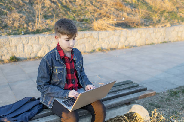 istock Cute boy is sitting on a bench near the river at sunset and using laptop to communicate with friends. Online education. 2117679552