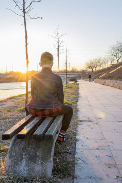 istock Cute boy is sitting on a bench near the river at sunset and using laptop to communicate with friends. Online education. 2117682174