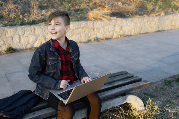 istock Cute boy is sitting on a bench near the river at sunset and using laptop to communicate with friends. Online education. 2117686095