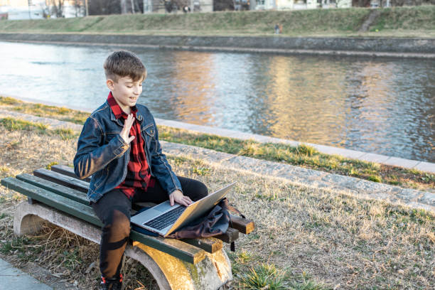 istock Cute boy is sitting on a bench near the river at sunset and using laptop to communicate with friends. Online education. 2117692428