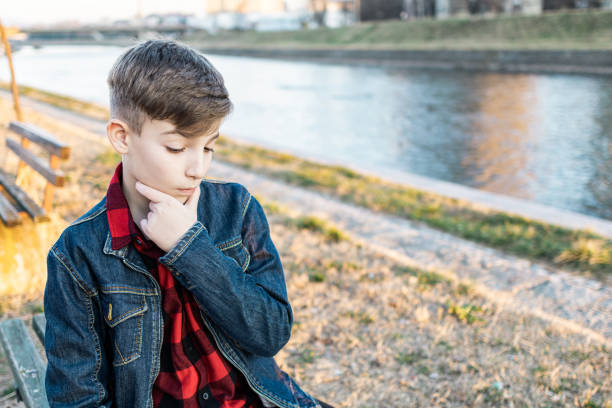 Cute boy is sitting on a bench near the river at sunset and using laptop to communicate with friends. Online education.