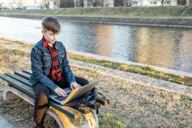 istock Cute boy is sitting on a bench near the river at sunset and using laptop to communicate with friends. Online education. 2117695732