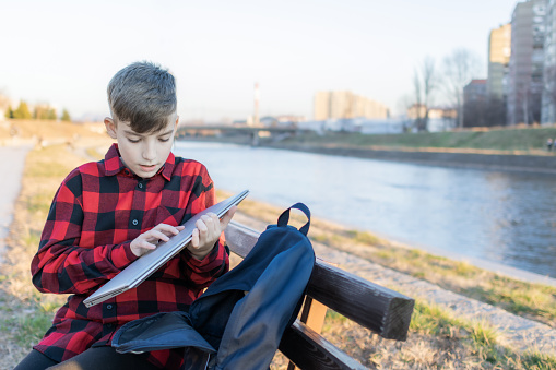 Cute boy is sitting on a bench near the river at sunset and using laptop to communicate with friends. Online education.