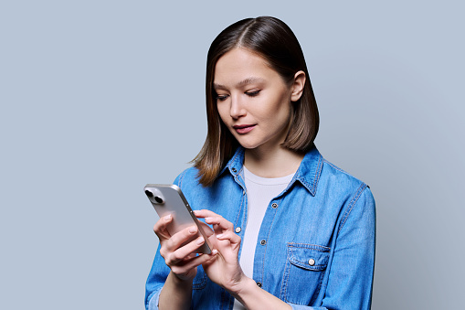 Young woman using smartphone in gray background. Serious 20s female looking at screen, texting. Mobile Internet applications apps, technologies for work education communication shopping healthcare