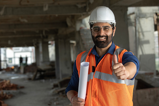 Portrait of confident smiling male building contractor holding blueprint and showing thumbs up sign at construction site