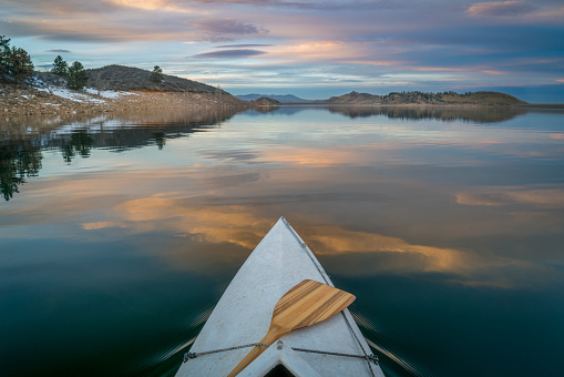 bow of expedition canoe with a wooden paddle at calm winter dusk over mountain lake - Horsetooth Reservoir