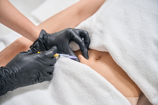 Partial image of female cosmetologist doing stomach beauty injection with syringe of woman on couch in beauty salon. Body skin care, lifting and rejuvenation