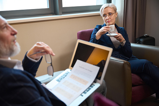 Adult caucasian businesswoman drinking tea or coffee and looking at thoughtful businessman with laptop and documents in hotel room at daytime. Business trip, vacation and travelling. Idea of teamwork