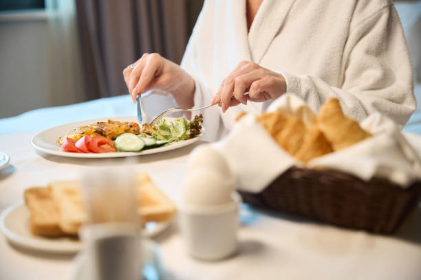 istock Cropped woman wearing bathrobe having breakfast at table in blurred hotel room 2117926229