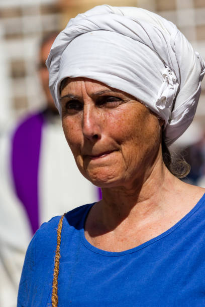 Close-up of the face of a woman. She wears a white cloak with a hood. She has her eyes closed and points to her open hand with her index finger.