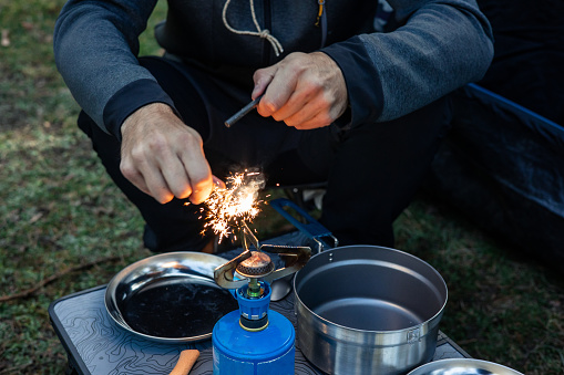 A camper is seen attempting to light a camping stove at the campsite in nature, with sparks flying from the flint, as he prepares to cook dinner amidst the wilderness