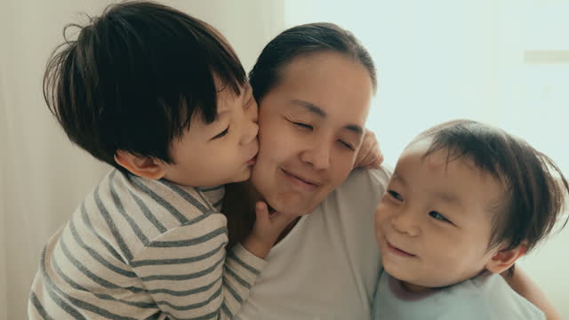 Asian Mother and Two Sons Embracing in Cozy Bedroom at Home, Enjoying Leisure Time Together with Genuine Smiles.