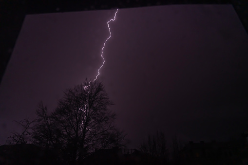 One lightning strike against purple sky on the tree at night, photo made through glass of the window.