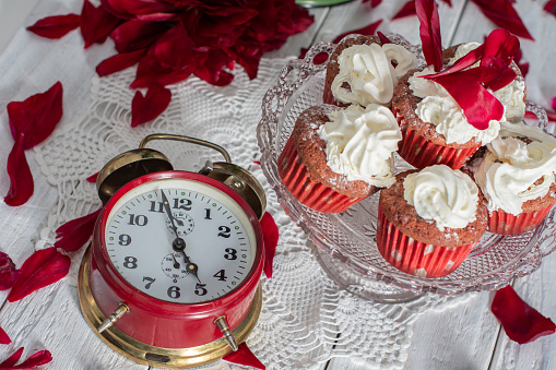 still life in English style with scarlet peonies and red velvet cupcakes with cream tops on a platter, red antique clock showing tea time,High quality photo