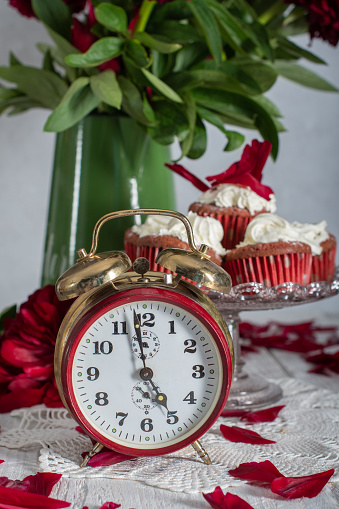 still life in English style with scarlet peonies and red velvet cupcakes with cream tops on a platter, red antique clock showing tea time,High quality photo