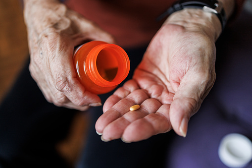 Close-up of an elderly woman's hands as she takes a single yellow pill from orange medicine bottle. Medication management for seniors