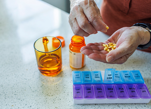 Close-up of Caucasian elderly woman's hands as she organizes her medication into daily pill organizer. Bottle of pills and glass of water resting on countertop