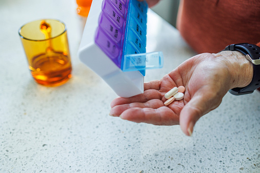 Senior woman prepares her daily medication, holding pills in her hand next to pill dispenser, with a glass of water on table. Close-up of elderly hand with medication, focus on hand with blurred background