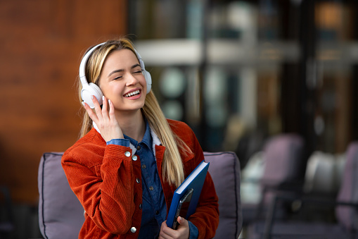 A joyful university student, wearing a bright smile, closes her eyes in bliss as she listens to music while flipping through an open book. Her radiant expression reflects the joy and relaxation found in the midst of study sessions. This image captures the harmonious blend of academic focus and personal enjoyment, portraying the student's ability to find moments of happiness amidst her studies. Perfect for showcasing the balance between work and leisure in university life in promotional materials and educational content.